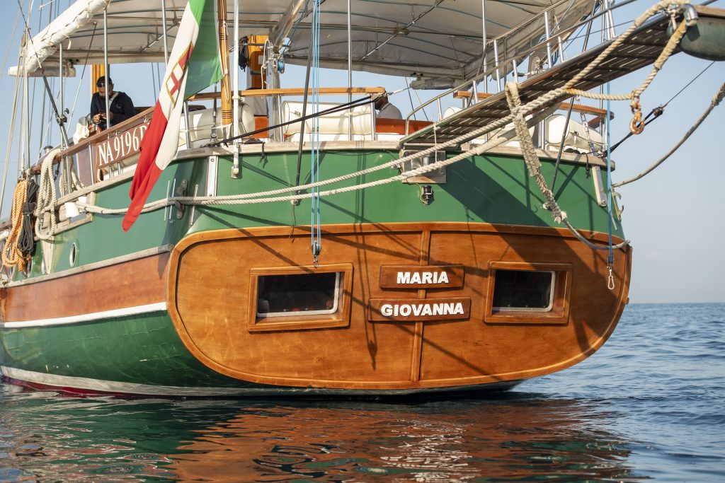The stern of a green and wooden sailboat named Maria Giovanna, with a red, white, and green flag, is shown floating on calm water under a clear sky.