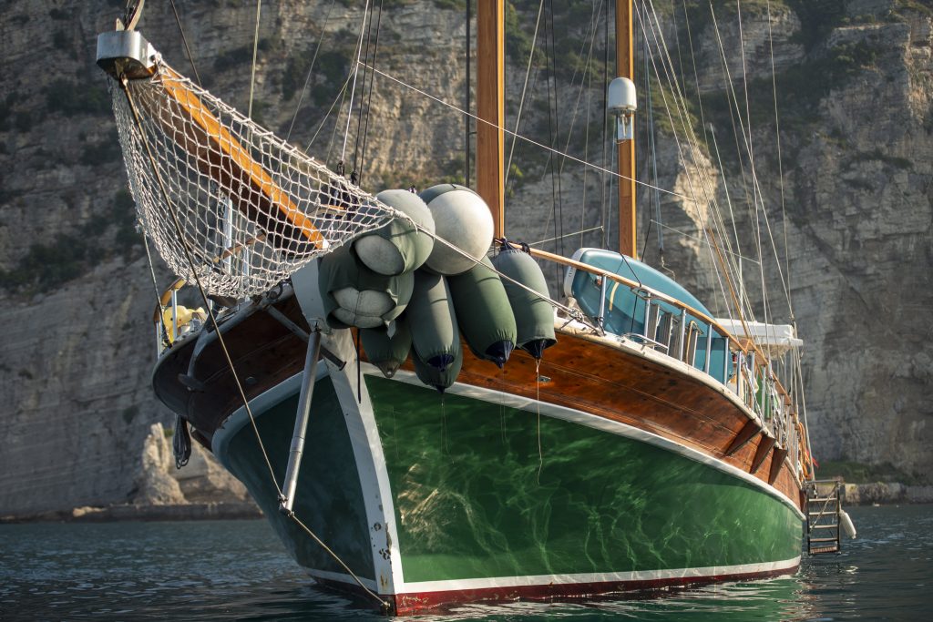 A large green and brown wooden sailboat with multiple bumpers tied to its side is anchored on calm water near a rocky cliff. The sunlight reflects on the boats hull and the cliffs are in the background.
