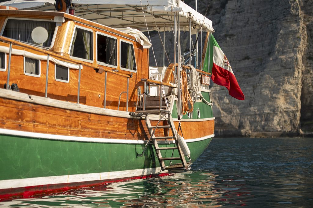 A wooden and green boat with an Italian flag is anchored on calm water near rocky cliffs. A metal ladder hangs off the boats side, and sunlight reflects off the water’s surface.