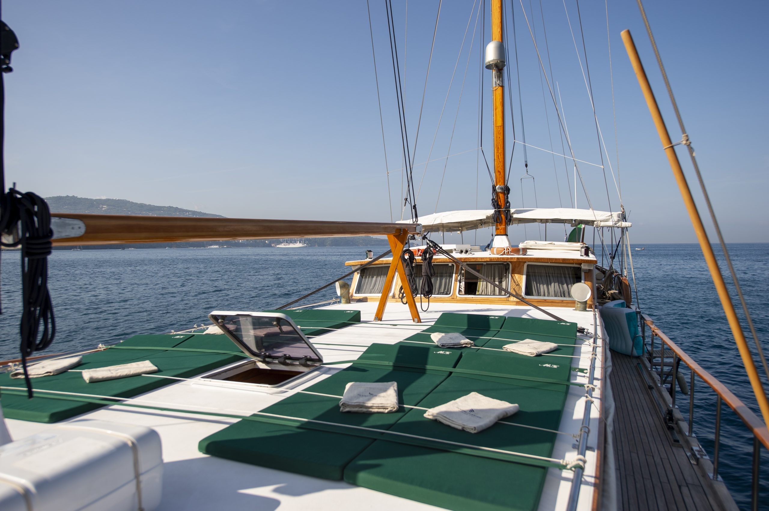 View from the deck of a yacht with green sun pads and a wooden mast, sailing on calm blue water near a distant coastline under a clear sky.