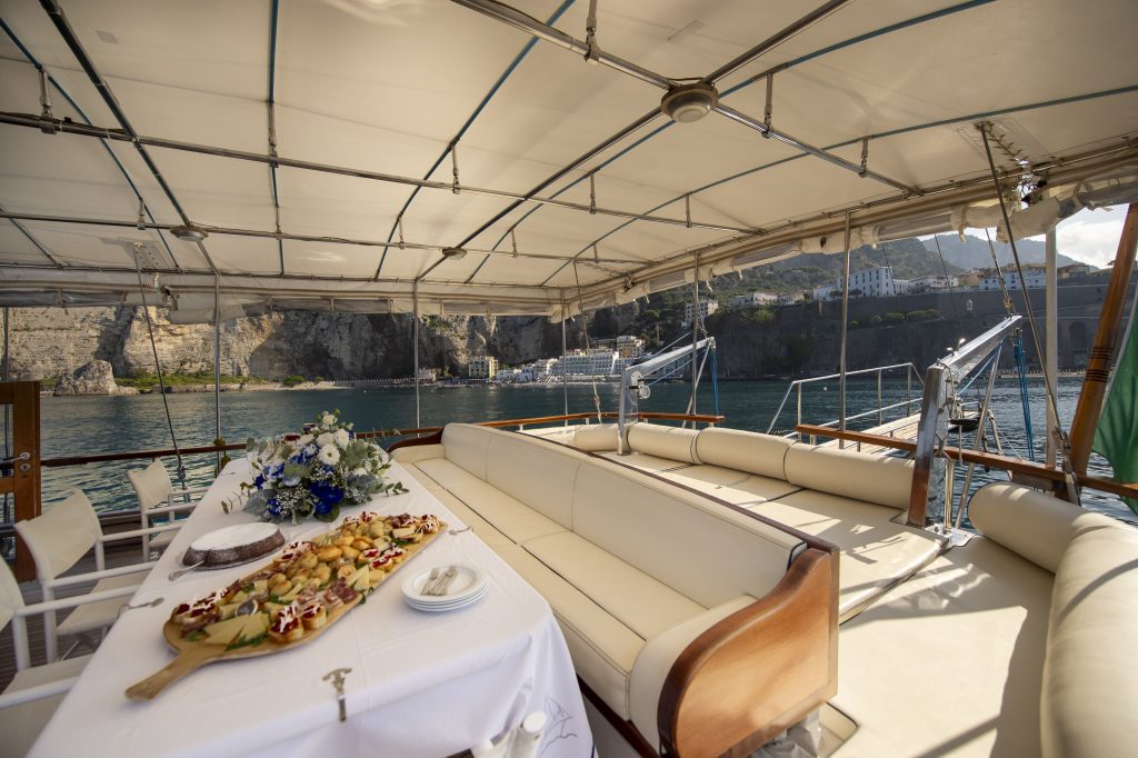 A luxurious boat deck with cream cushions, a dining table set with plates, a floral centerpiece, and a platter of appetizers, overlooking calm blue water and rocky cliffs with buildings in the background.