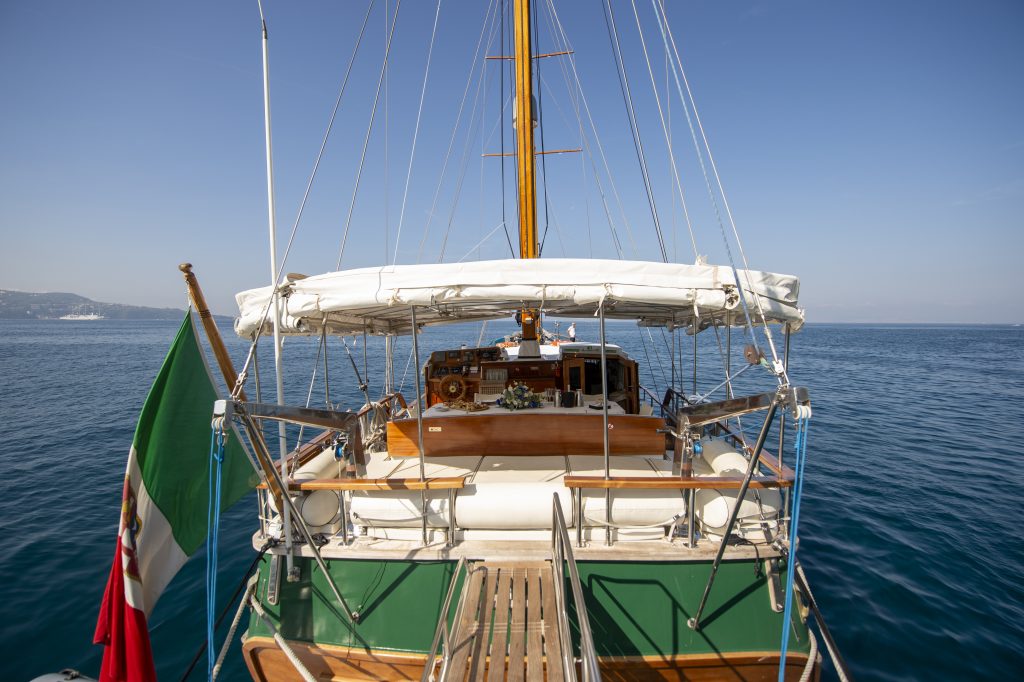 A view from the stern of a sailboat with a wooden deck, green accents, and an Italian flag, floating on calm blue water under a clear sky. The cabin and mast are visible at the center.
