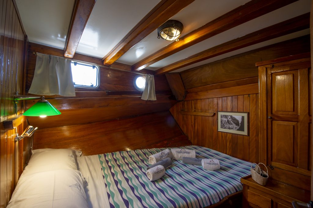 A cozy wooden cabin room with a bed covered in a striped blanket, towels neatly arranged on top, a small window, green lamp, framed photo on the wall, and built-in wood cabinets.