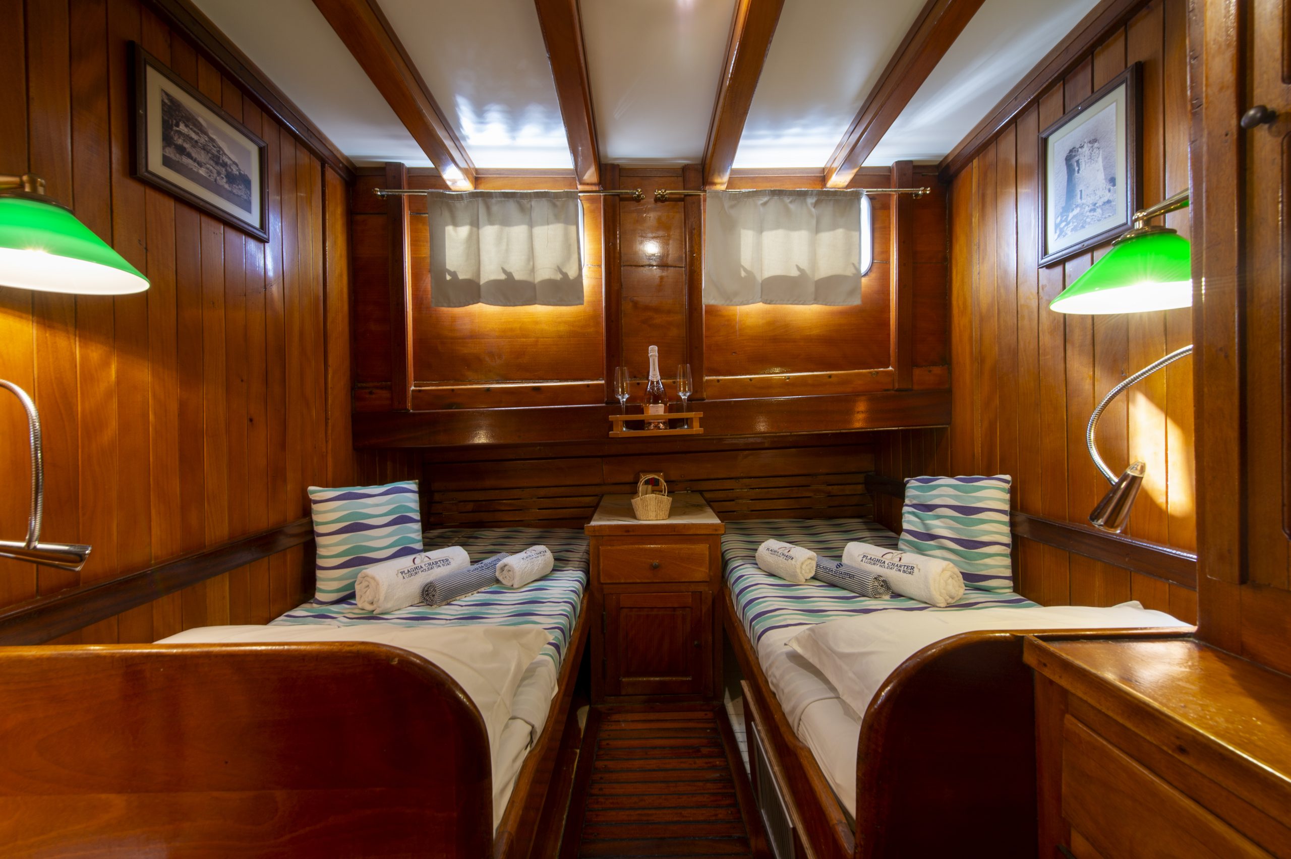Cozy wooden ship cabin with two single beds, white sheets, striped pillows, rolled towels, green lamps, framed pictures on the walls, and light streaming through small curtained windows.