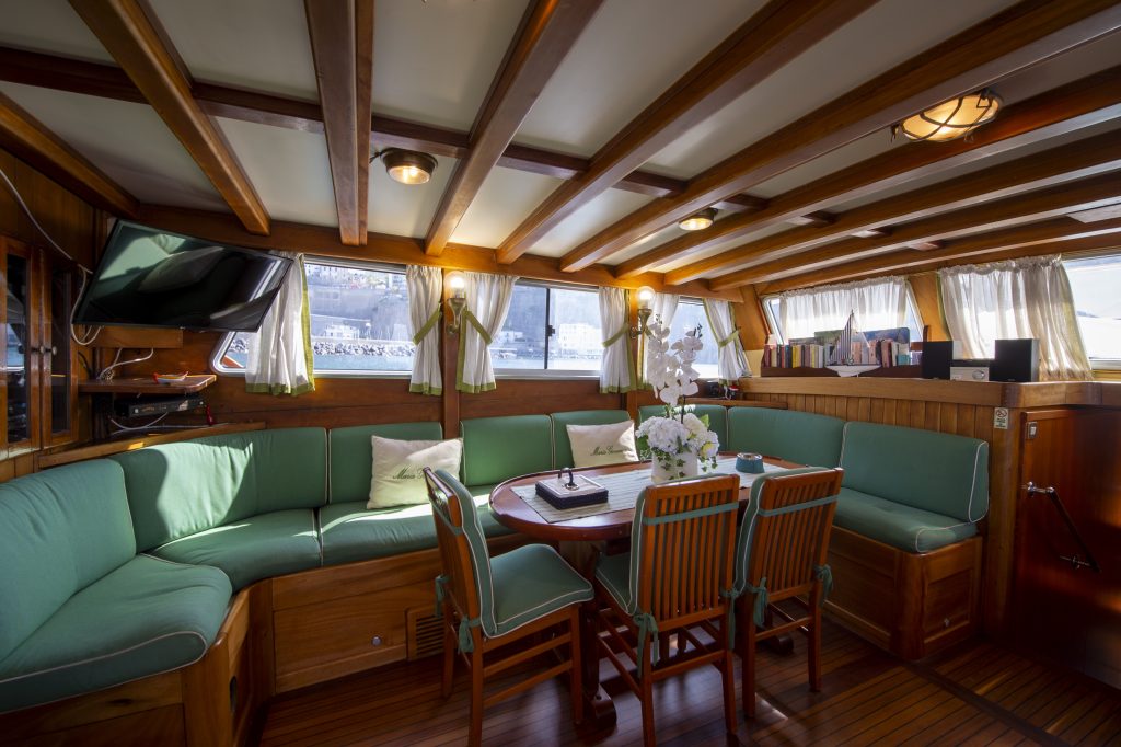 The interior of a boat with wooden beams, green cushioned seating, a round table with four wooden chairs, large windows with white curtains, and sunlight streaming inside.