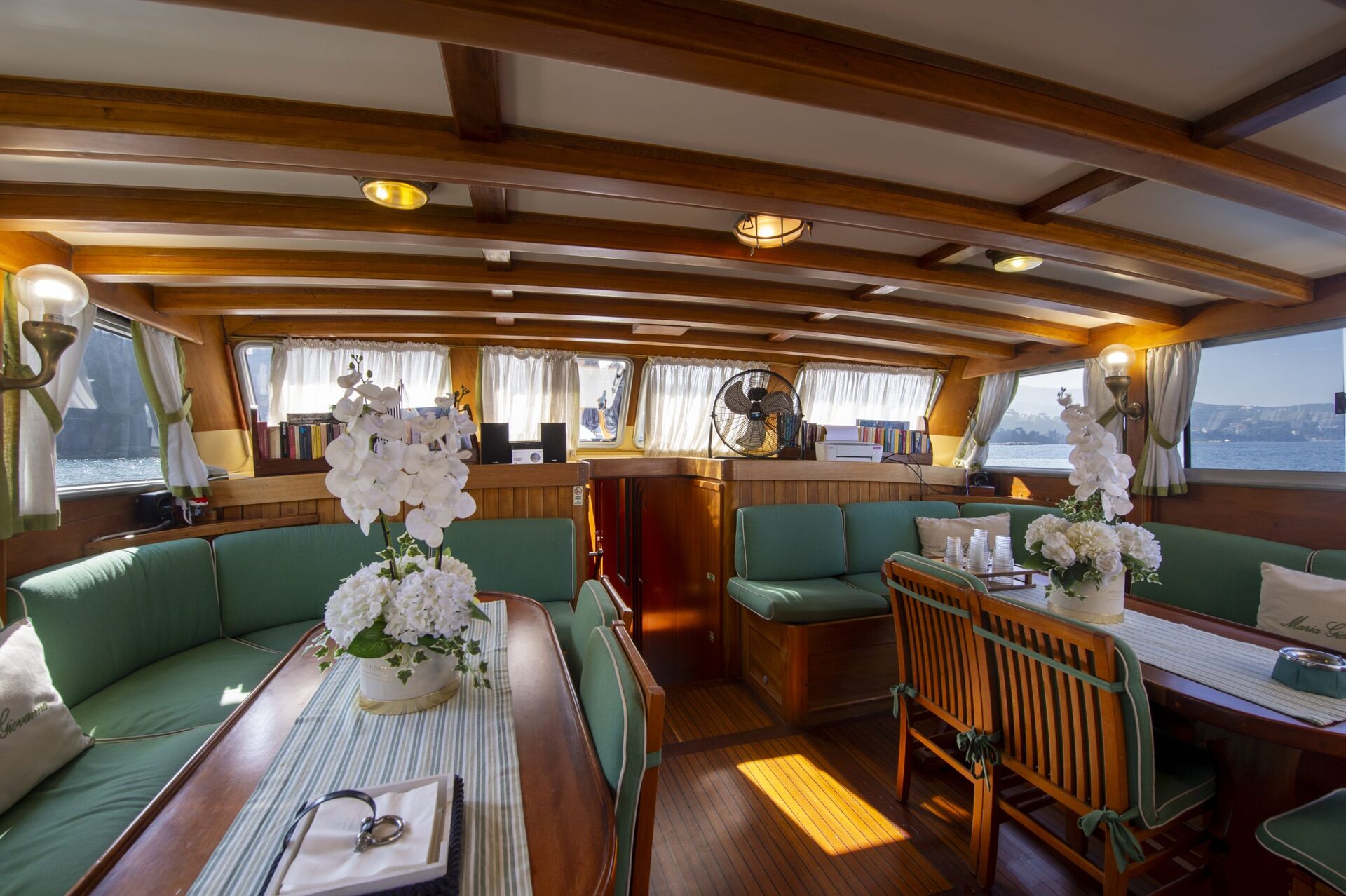 The cozy interior of a boat with wooden beams, green cushioned seating, a dining table with chairs, white flower arrangements, soft lighting, and large windows with curtains letting in daylight.