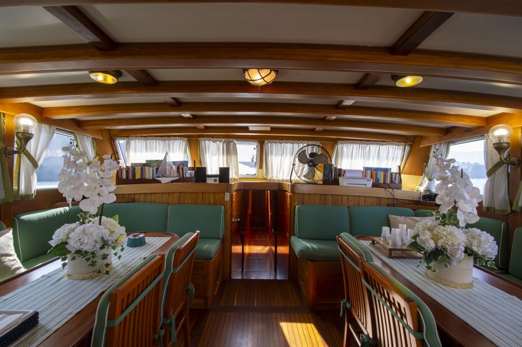 The interior of a boat with wooden beams, green cushioned seating, and tables decorated with white flowers. Sunlight streams through curtained windows, and books and a fan are on a rear shelf.