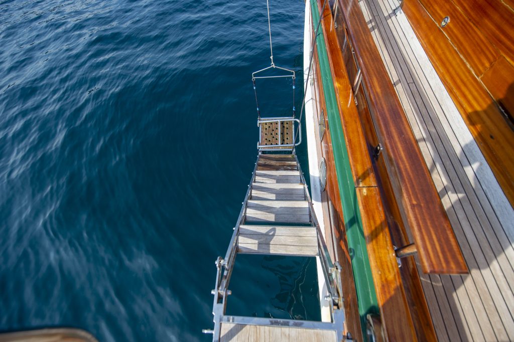 A wooden gangplank extends from the deck of a boat down to the calm blue sea, casting shadows on the water below. The boat’s polished wooden deck is visible along the right side of the image.
