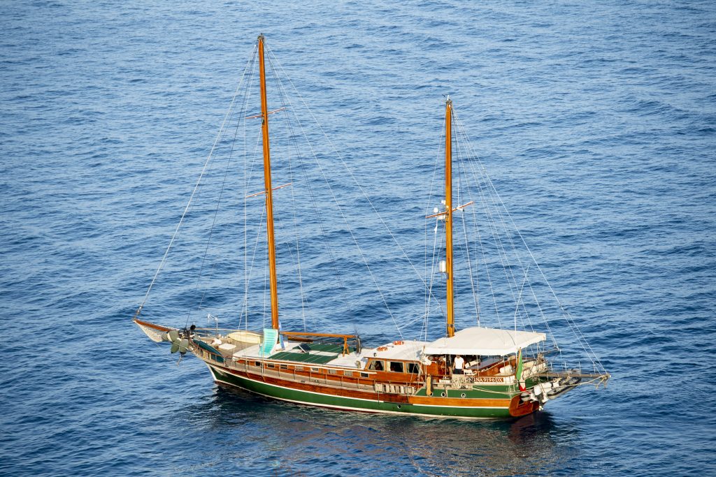 A wooden sailboat with two masts floats on calm, deep blue water. Several people are on board under a white canopy, and the boat has green, brown, and white coloring.