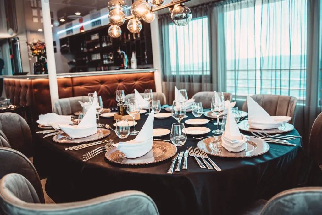 A round dining table set for eight with folded napkins, wine glasses, plates, and cutlery in an elegant restaurant with modern decor and large windows letting in natural light.