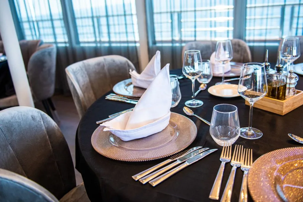 A round dining table set with elegant glassware, plates, silverware, and neatly folded white napkins in a bright restaurant with gray upholstered chairs.