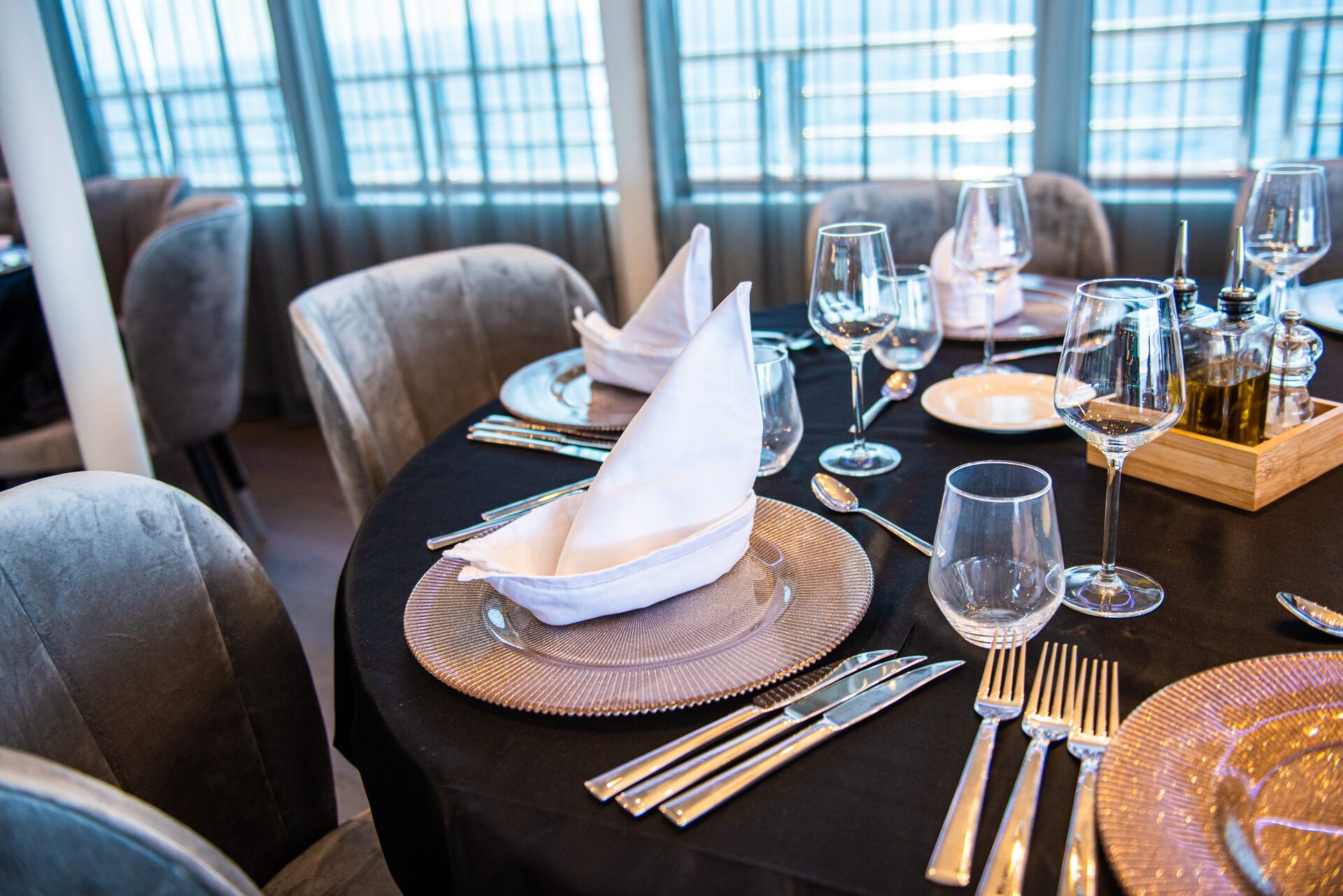 A round dining table set with elegant glassware, plates, silverware, and neatly folded white napkins in a bright restaurant with gray upholstered chairs.