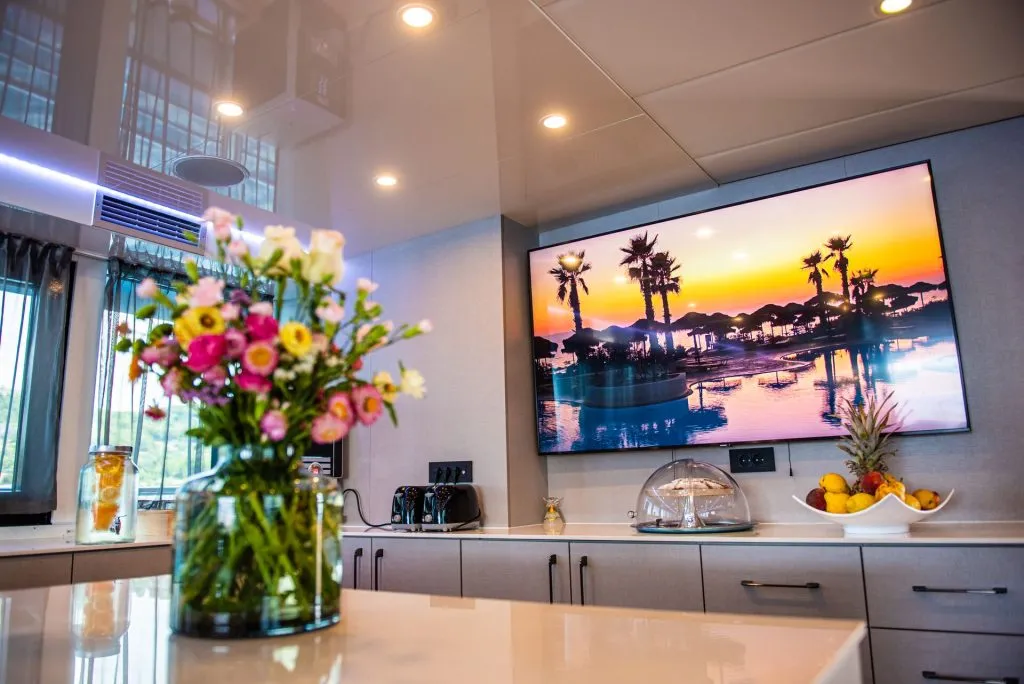 A modern kitchen with a vase of colorful flowers on the counter, a bowl of fruit, and a large wall-mounted TV displaying a tropical sunset scene with palm trees and water.