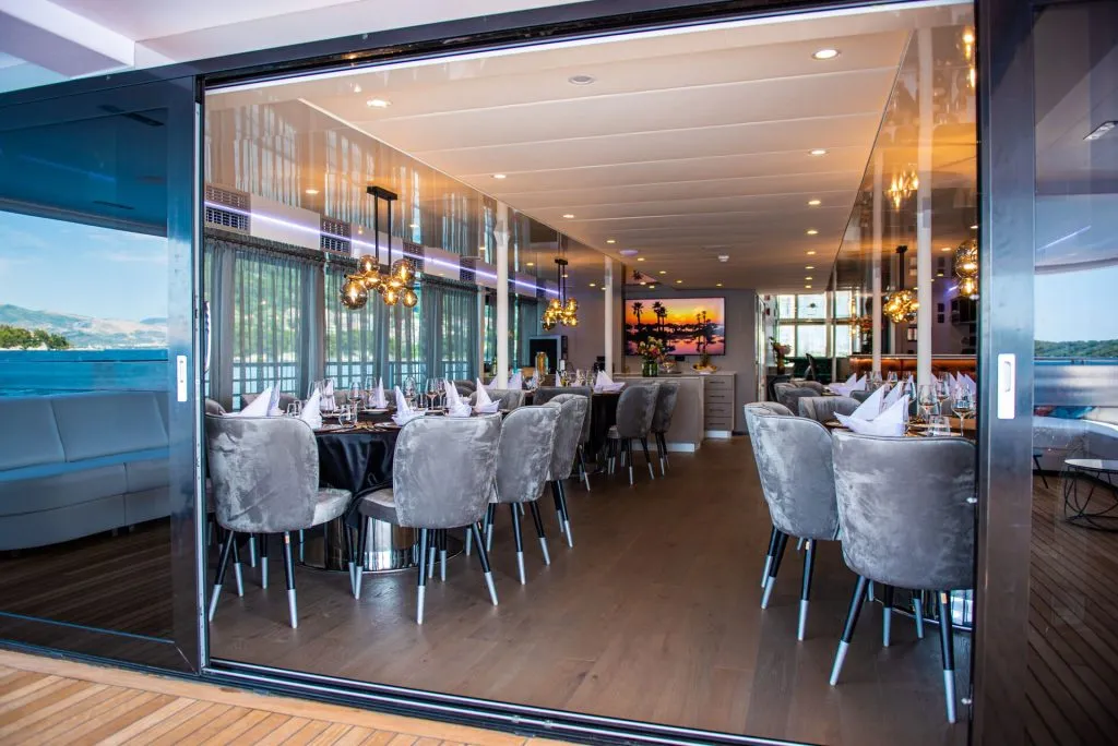 Elegant dining room on a boat with grey velvet chairs, set tables with white napkins, modern chandeliers, wood flooring, and large glass doors offering a view of the water and surrounding landscape.