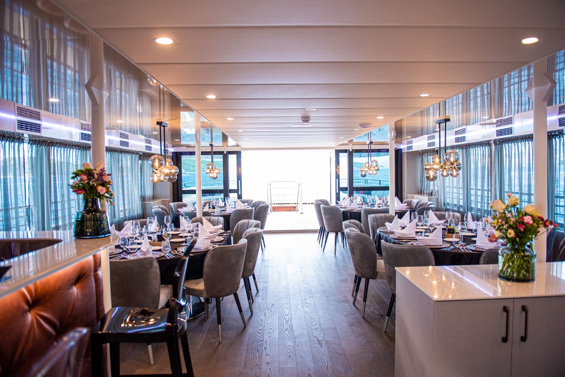 Elegant dining room with large windows, set tables with white linens and glasses, plush gray chairs, modern pendant lights, and floral centerpieces, looking out to a bright deck area at the far end.
