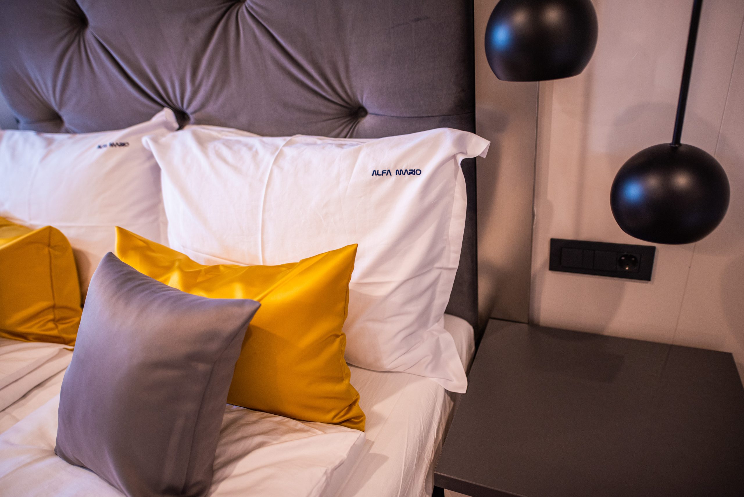 A modern bed with white and yellow pillows, a tufted gray headboard, and a gray nightstand beside it, with two black spherical pendant lights hanging above.
