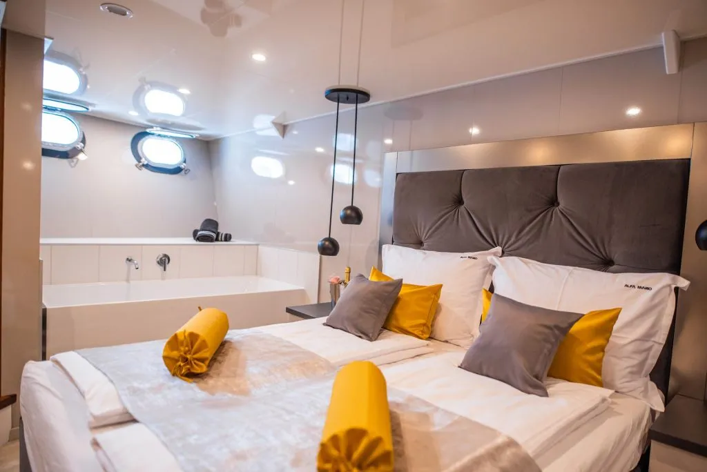 A modern bedroom with a large bed, gray and yellow pillows, and rolled yellow blankets. There is a bathtub in the background, with towels and small circular windows above it, creating a cozy and luxurious ambiance.