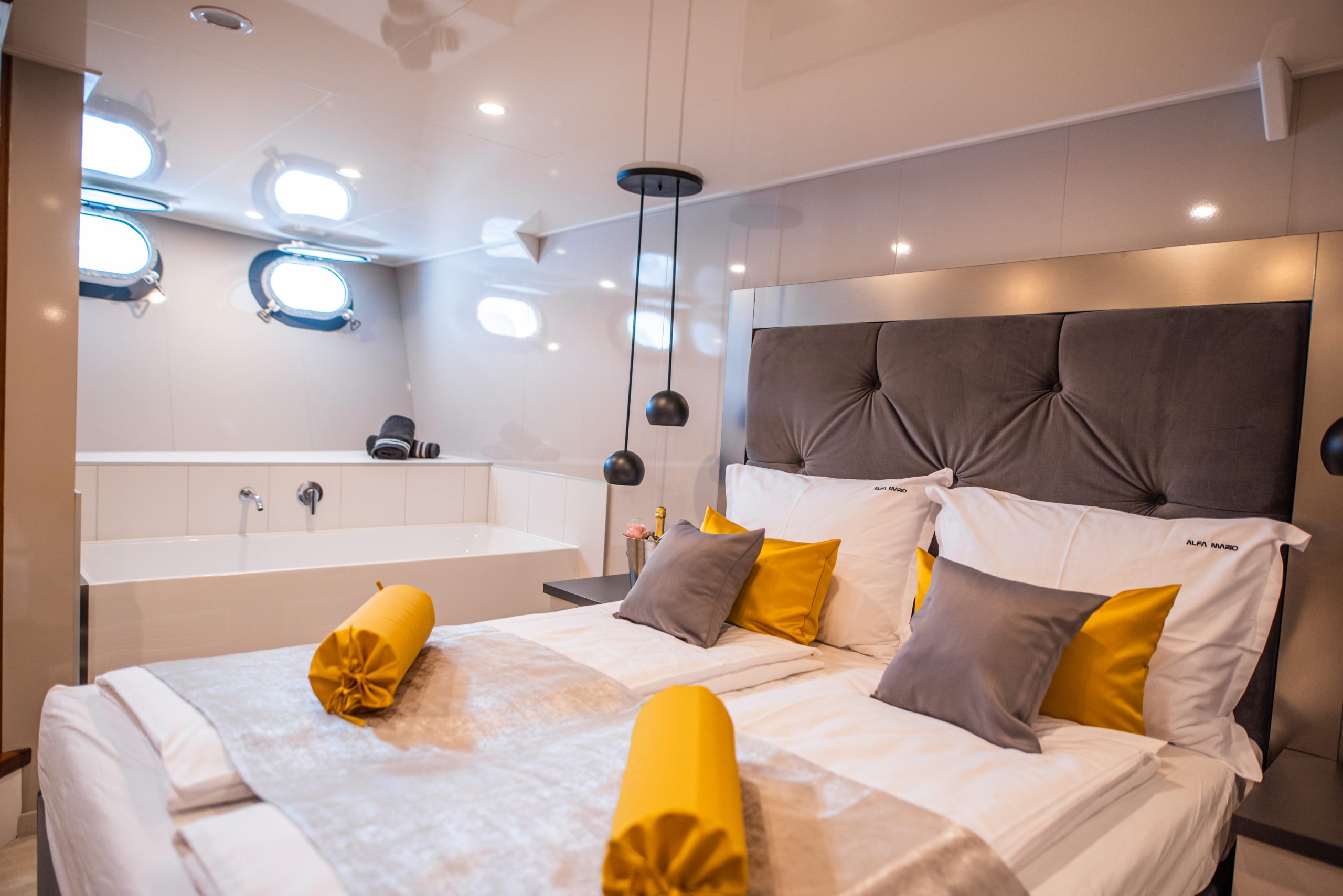 A modern bedroom with a large bed, gray and yellow pillows, and rolled yellow blankets. There is a bathtub in the background, with towels and small circular windows above it, creating a cozy and luxurious ambiance.