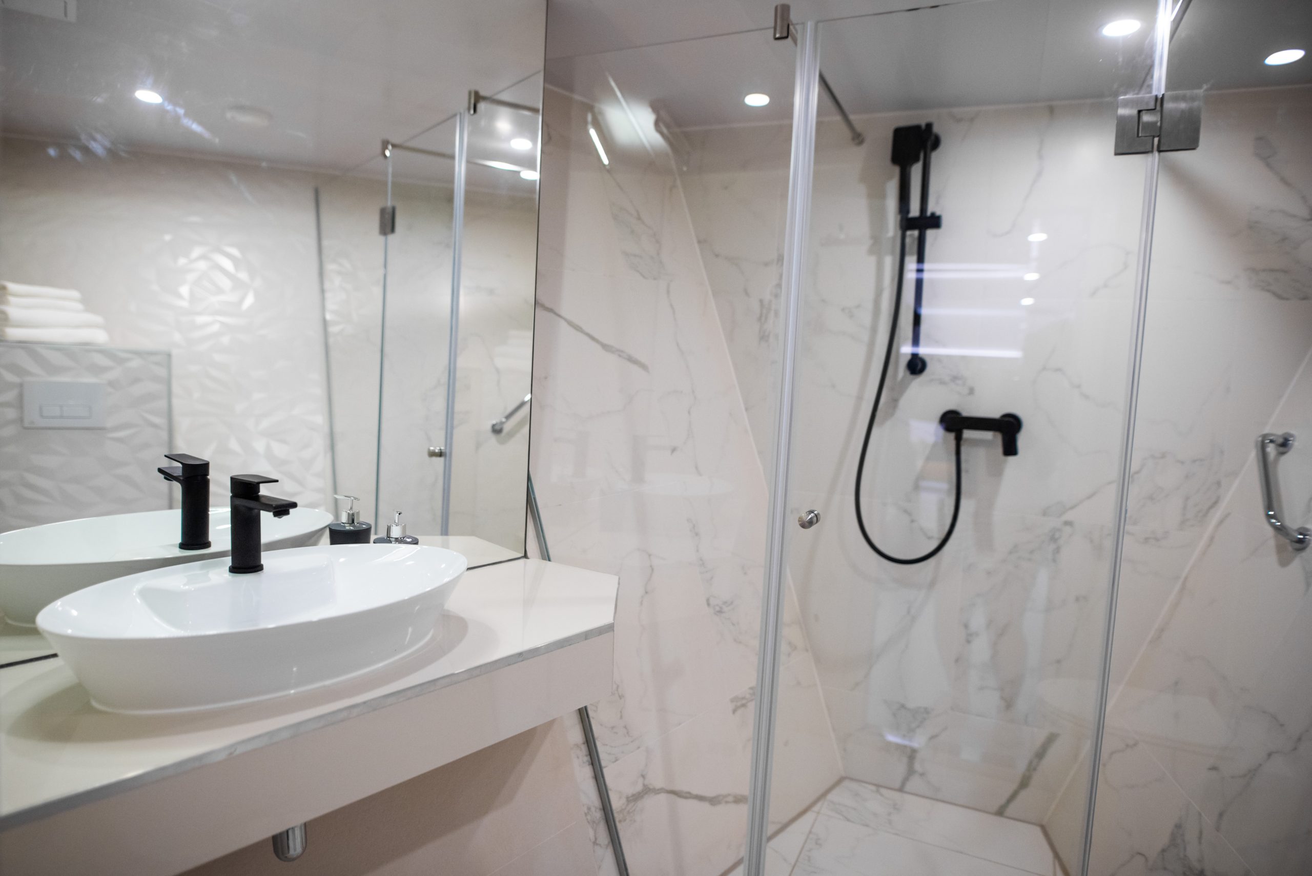 Modern bathroom with a glass-enclosed shower, marble-patterned walls, a white sink with black fixtures, towels stacked on a shelf, and a large mirror reflecting the room.