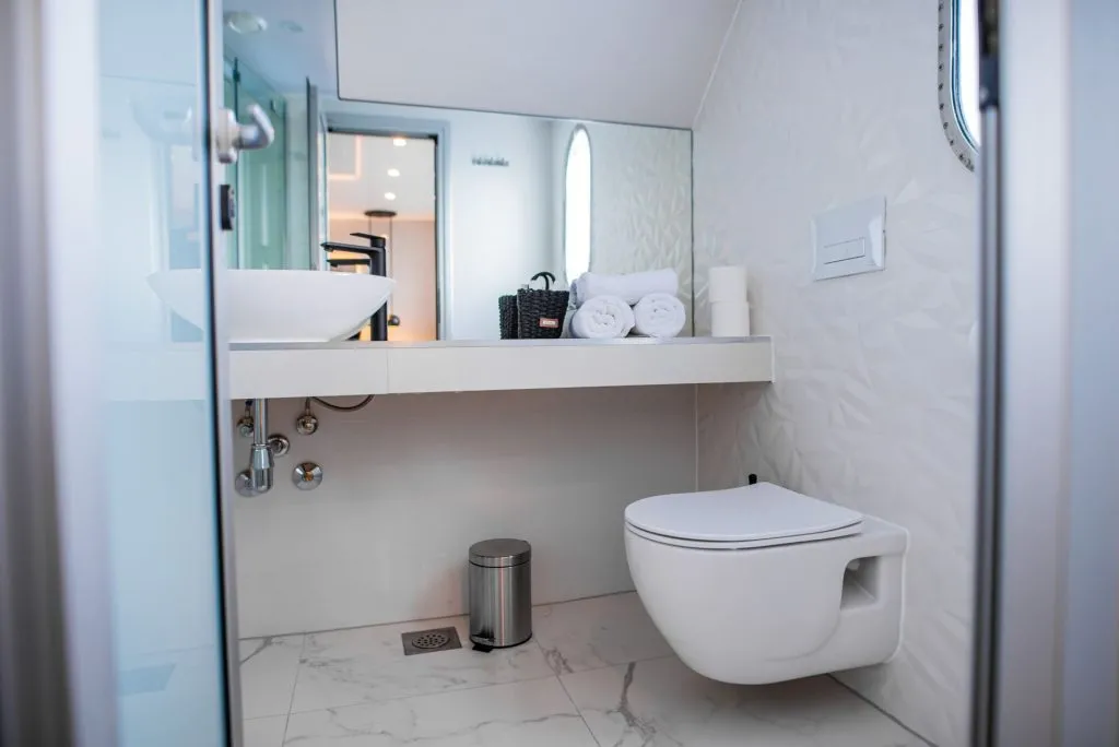 Modern bathroom with a floating toilet, white vessel sink, rolled towels, and toiletries on a counter. The design features sleek lines, white walls with subtle patterns, and marble-like flooring.