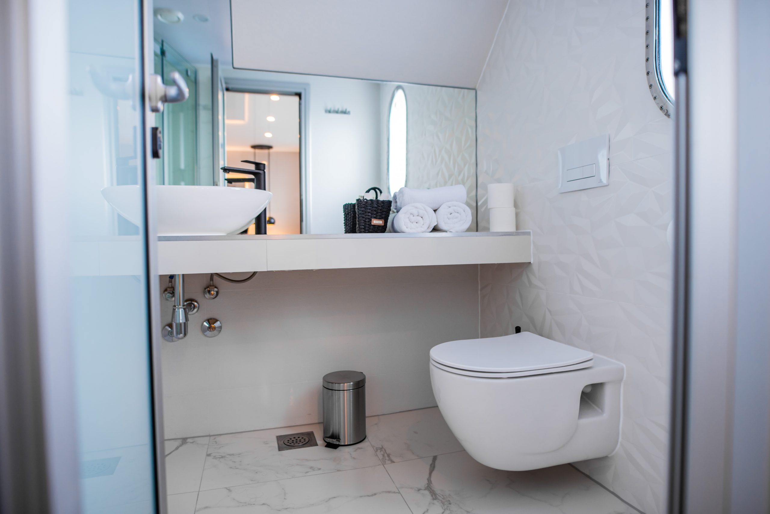 Modern bathroom with a floating toilet, white vessel sink, rolled towels, and toiletries on a counter. The design features sleek lines, white walls with subtle patterns, and marble-like flooring.