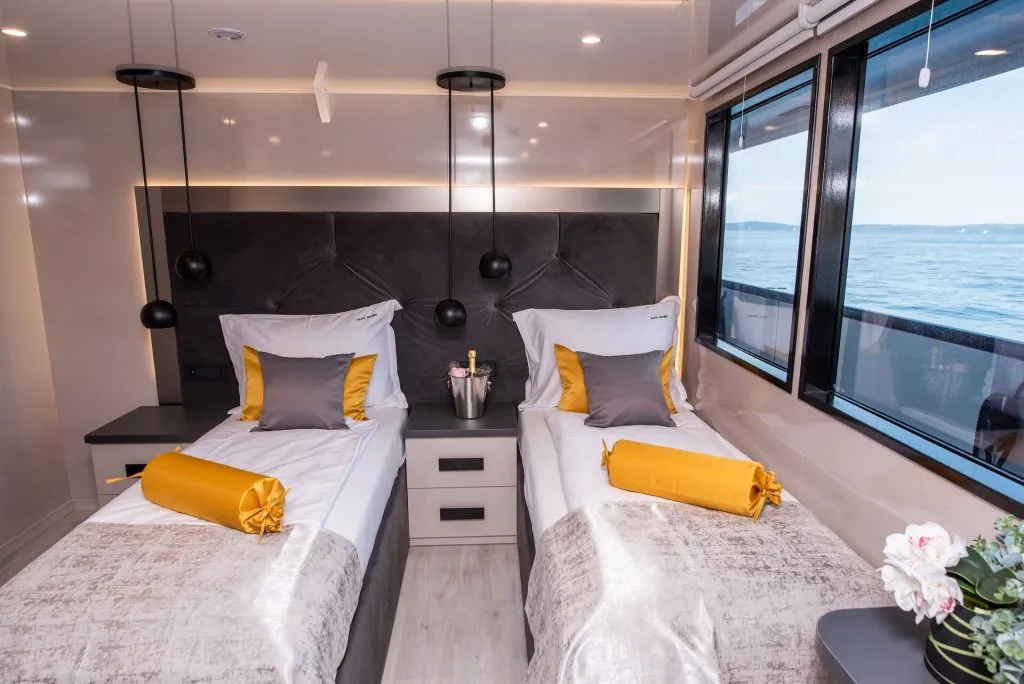 A modern cabin with two single beds, each with white, gray, and gold bedding. There are hanging pendant lights, a window with a sea view, a bedside table with a champagne bucket, and a vase of flowers.