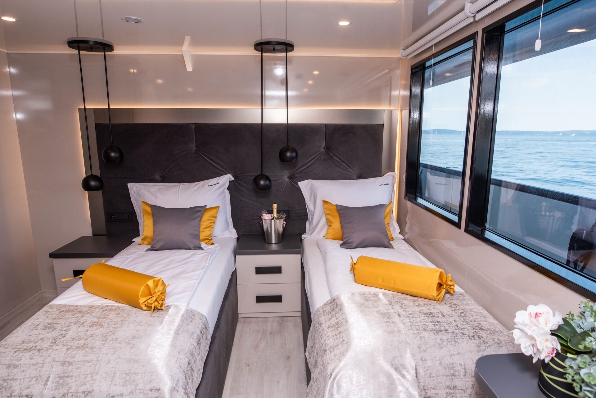 A modern cabin with two single beds, each with white, gray, and gold bedding. There are hanging pendant lights, a window with a sea view, a bedside table with a champagne bucket, and a vase of flowers.
