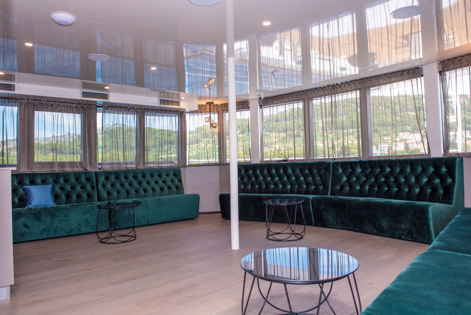 Spacious lounge with green velvet tufted sofas, round glass coffee tables, large windows with sheer curtains, and views of greenery outside. The room has light wooden floors and a modern, airy atmosphere.