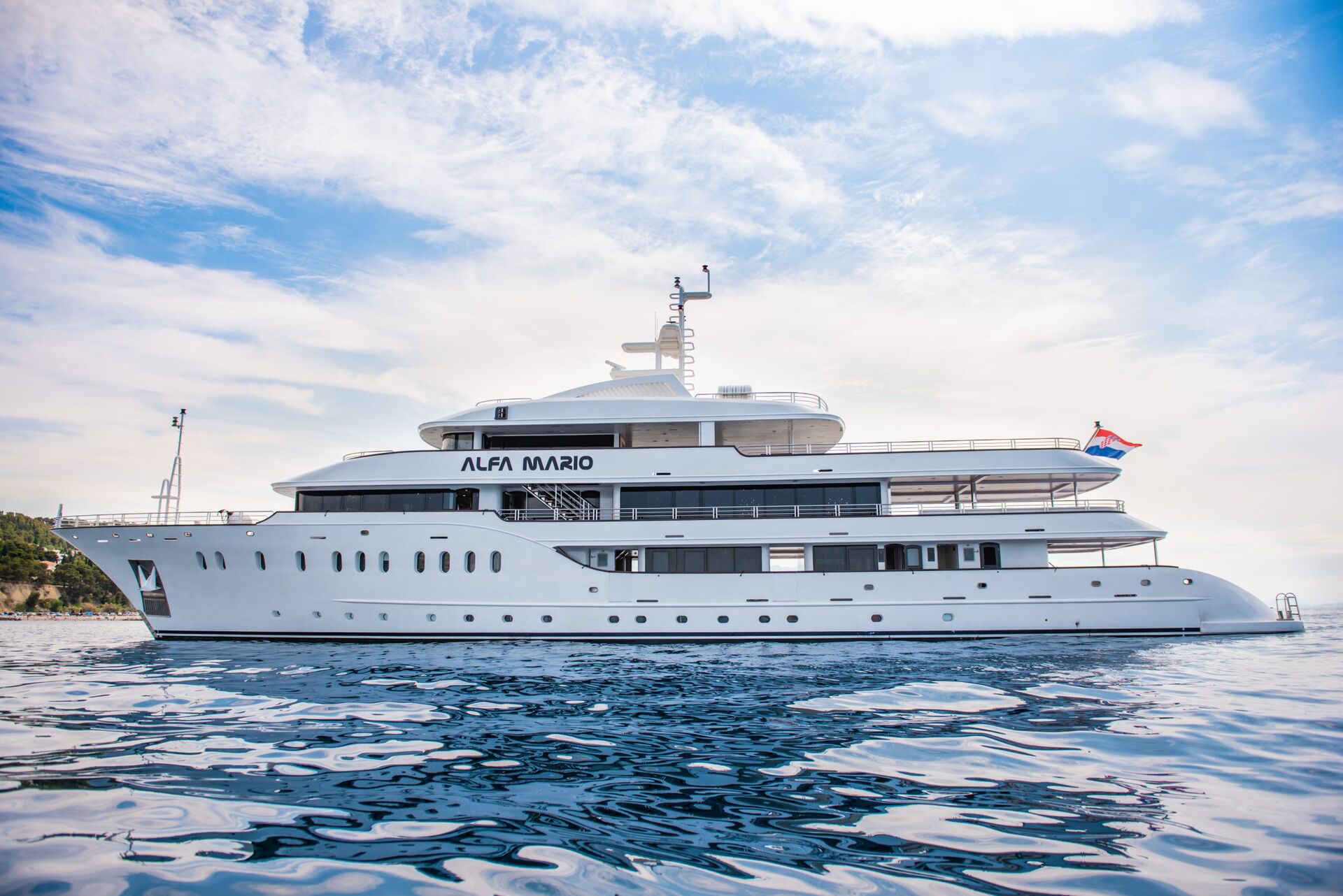 A large white luxury yacht named Alfa Mario is anchored on calm blue water under a partly cloudy sky, with a distant shoreline visible on the left. A flag is flying at the stern of the yacht.