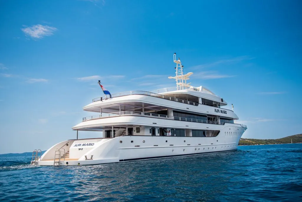 A large white luxury yacht named Aura Marco sails on calm blue water under a clear sky, with land visible in the background.