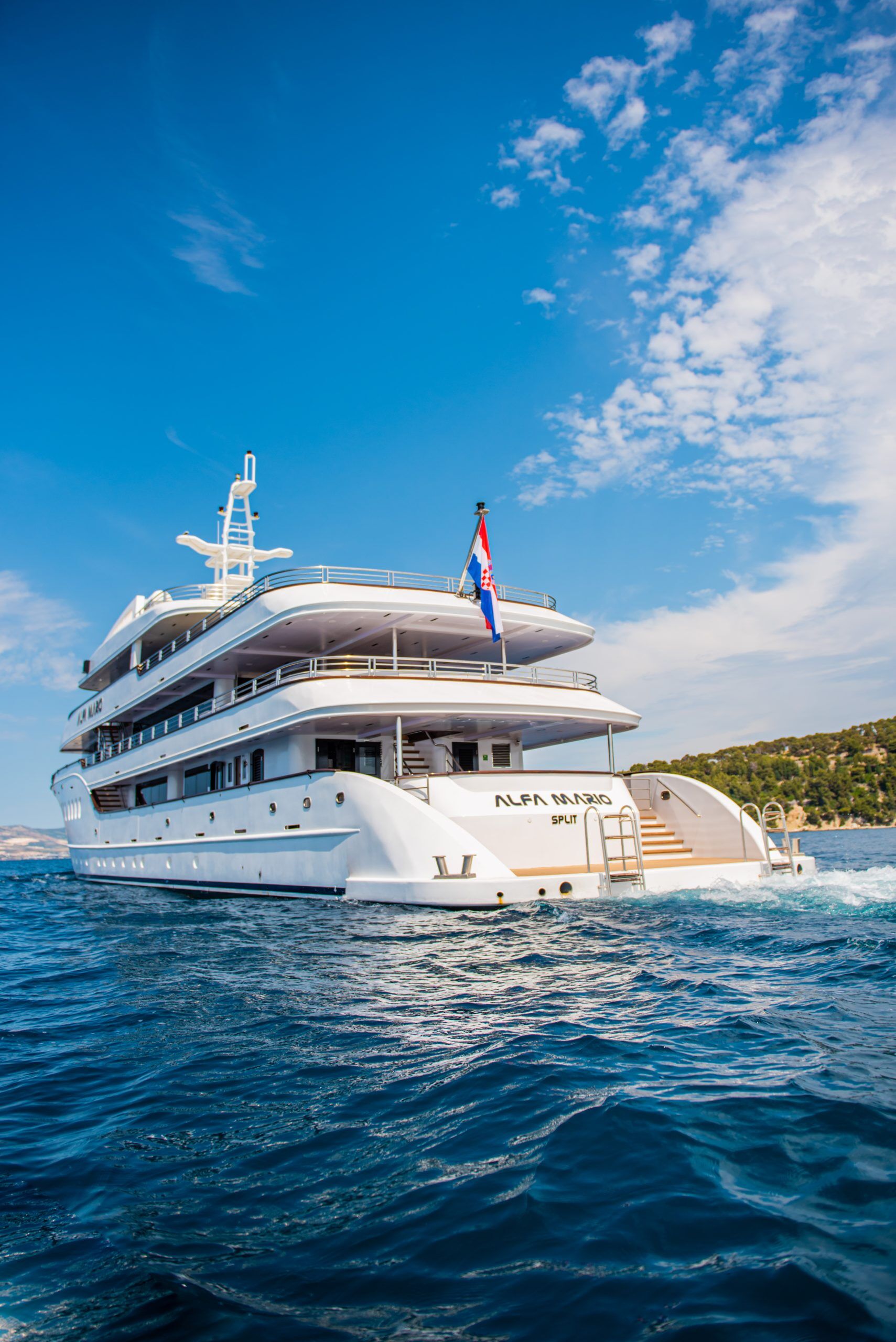 A large white luxury yacht named Alfa Mario sails on clear blue water under a partly cloudy sky, with a Croatian flag flying at the stern and a forested coastline in the background.