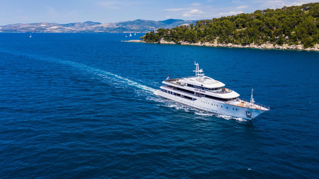 A large white yacht sails through bright blue water near a green, tree-covered coastline, with distant mountains and a few small sailboats visible under a clear sky.