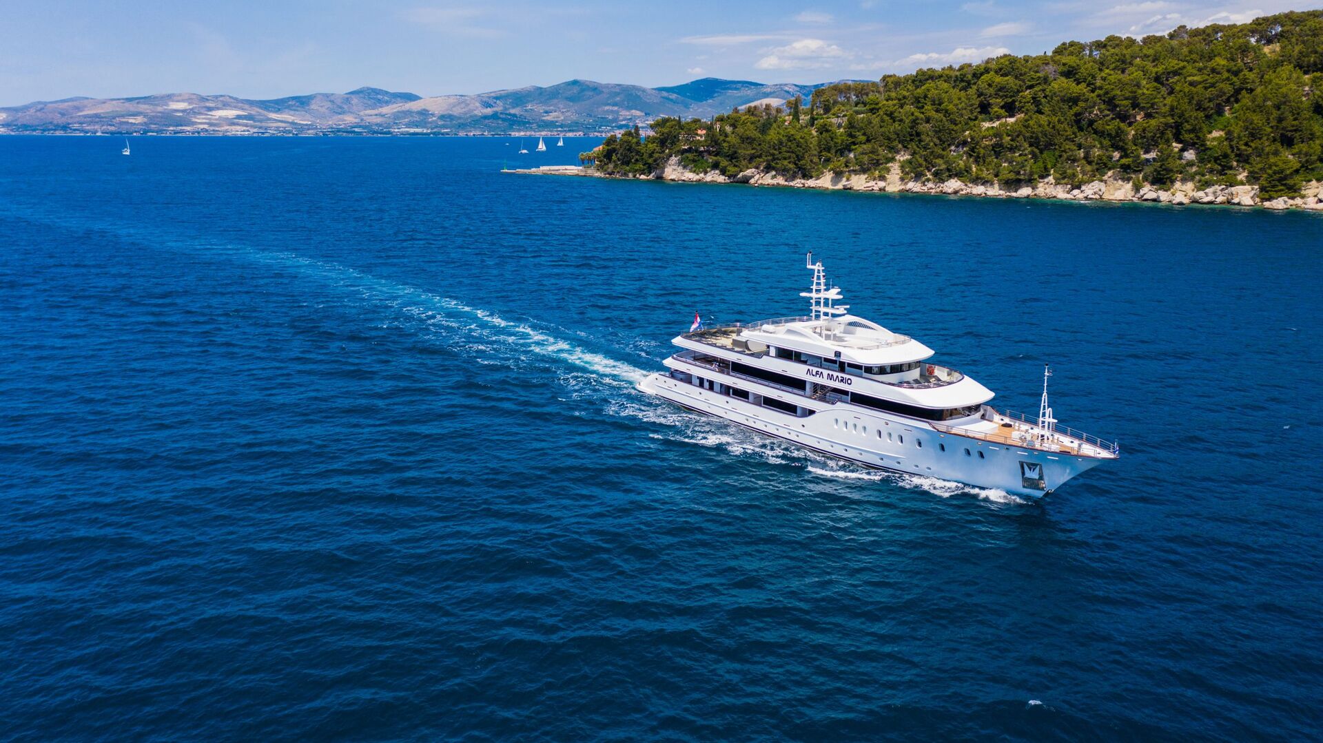 A large white yacht sails through bright blue water near a green, tree-covered coastline, with distant mountains and a few small sailboats visible under a clear sky.