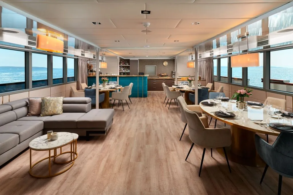 A spacious, modern dining and lounge area with round tables, plush chairs, a sectional sofa, and large windows offering ocean views. Warm lighting and elegant décor create a luxurious, inviting atmosphere.