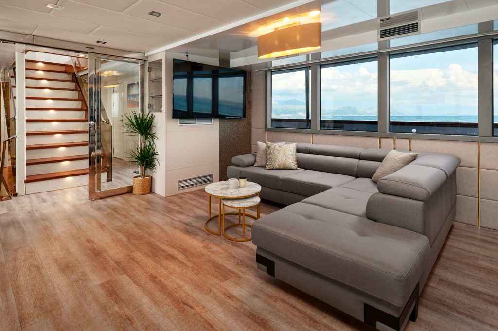 Modern living area with large gray sectional sofa, round coffee tables, potted plant, wall-mounted TV, wooden staircase, and large windows offering a view of the sea and sky.