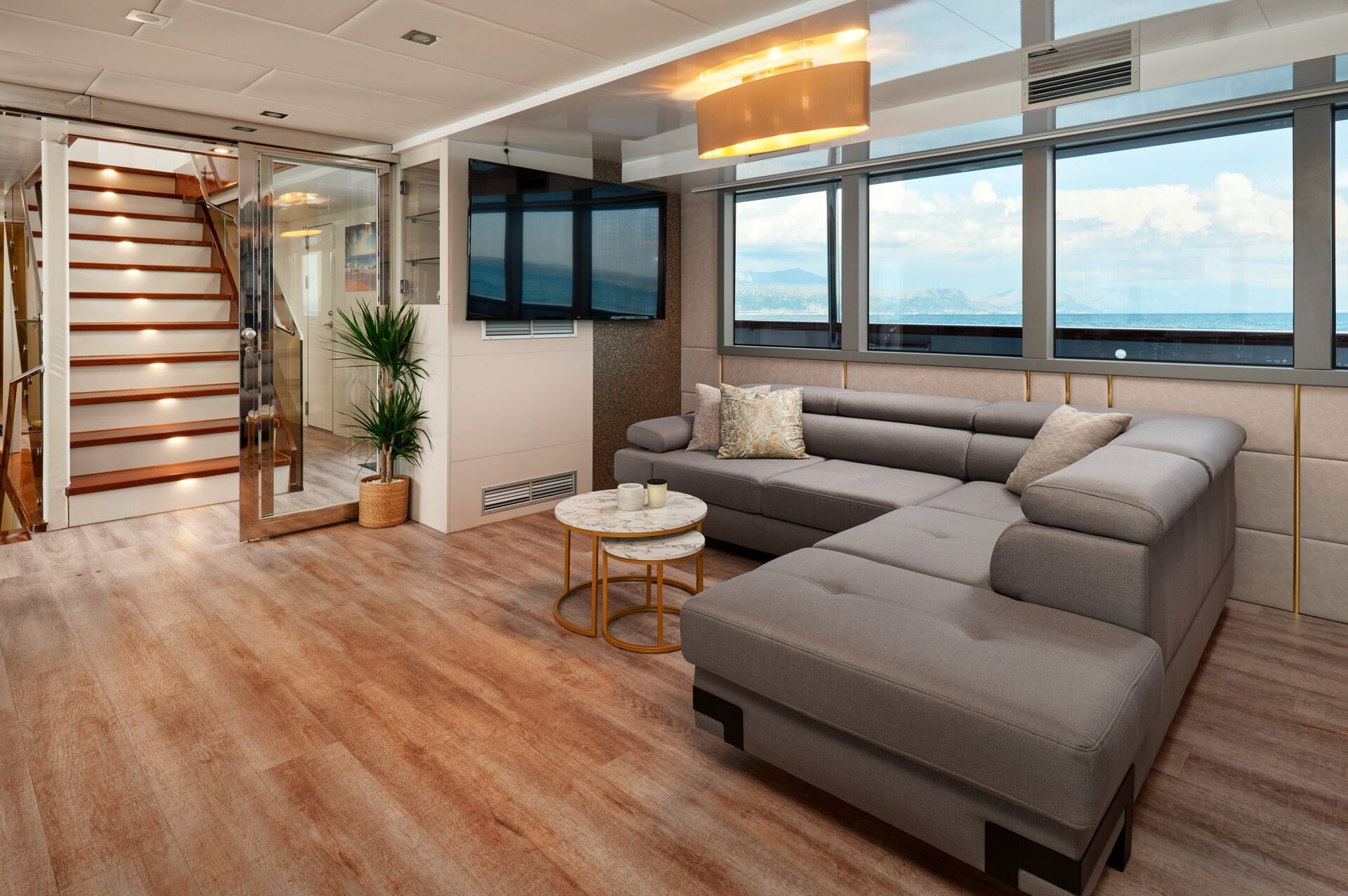 Modern living area with large gray sectional sofa, round coffee tables, potted plant, wall-mounted TV, wooden staircase, and large windows offering a view of the sea and sky.