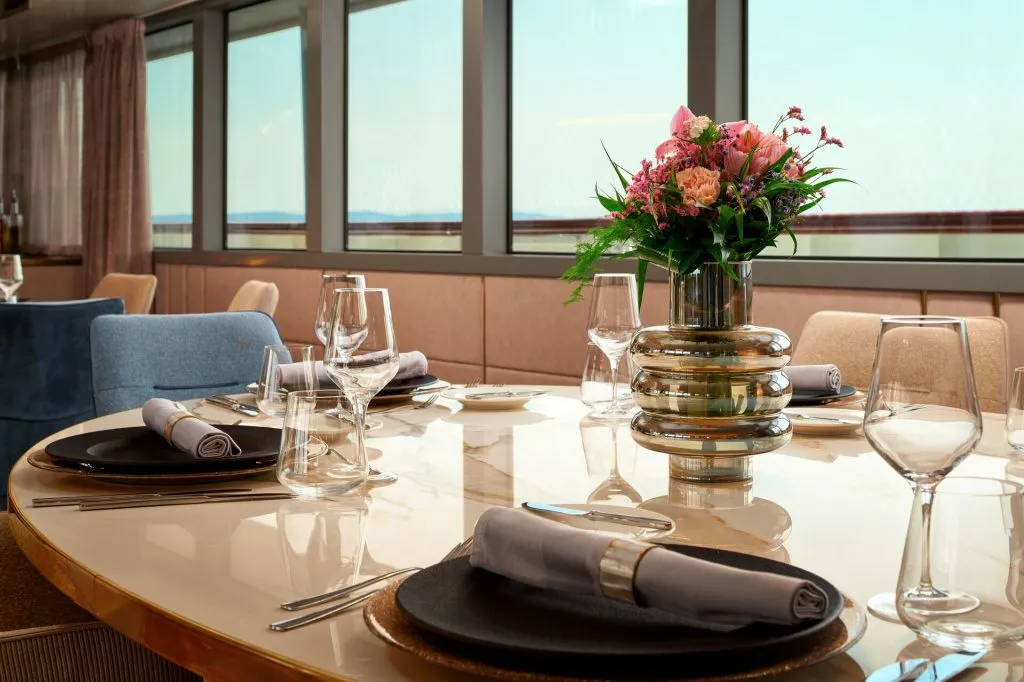A modern, elegant dining table set with plates, glasses, napkins, and a metallic vase of pink and purple flowers, positioned by large windows with a scenic outdoor view.