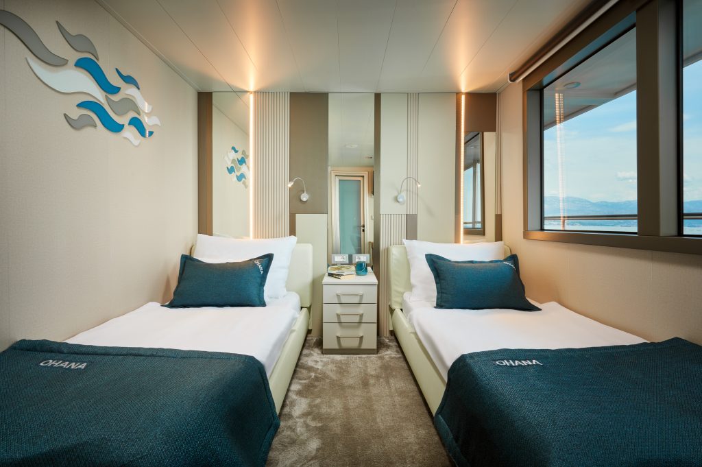 A modern cabin with two single beds, teal bedding, white walls, wall art, reading lamps, a window with a water view, and a small nightstand between the beds.