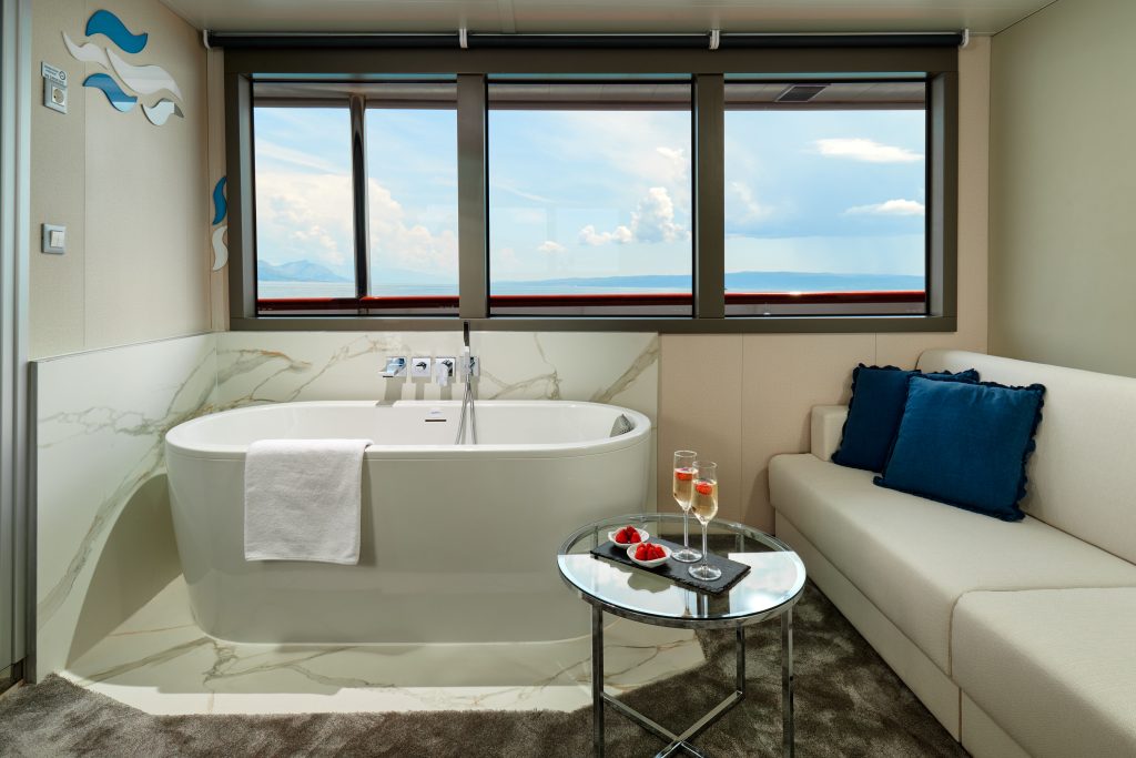 A modern bathroom with a freestanding white bathtub, a towel draped over the side, a glass table with drinks and strawberries, a white sofa with blue pillows, and large windows showing a scenic sky view.