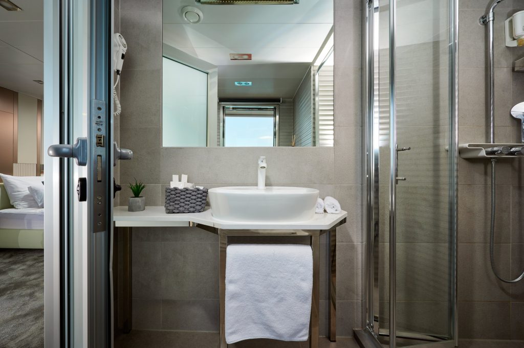 Modern bathroom with a white sink on a countertop, a towel hanging below, a grey basket with toiletries, a potted plant, and a glass-enclosed shower. A glimpse of a bedroom is visible through the open door.