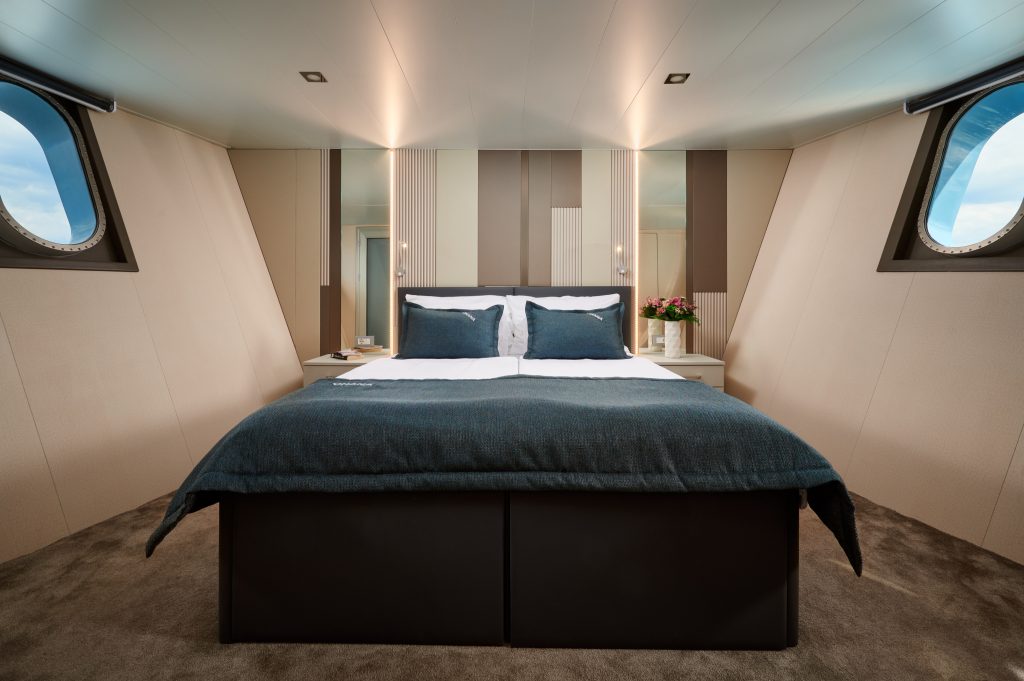 Modern bedroom with a large bed, blue bedding, neutral striped accent wall, round windows on each side, bedside tables, and a vase of flowers on the right. The room has a cozy and luxurious feel.