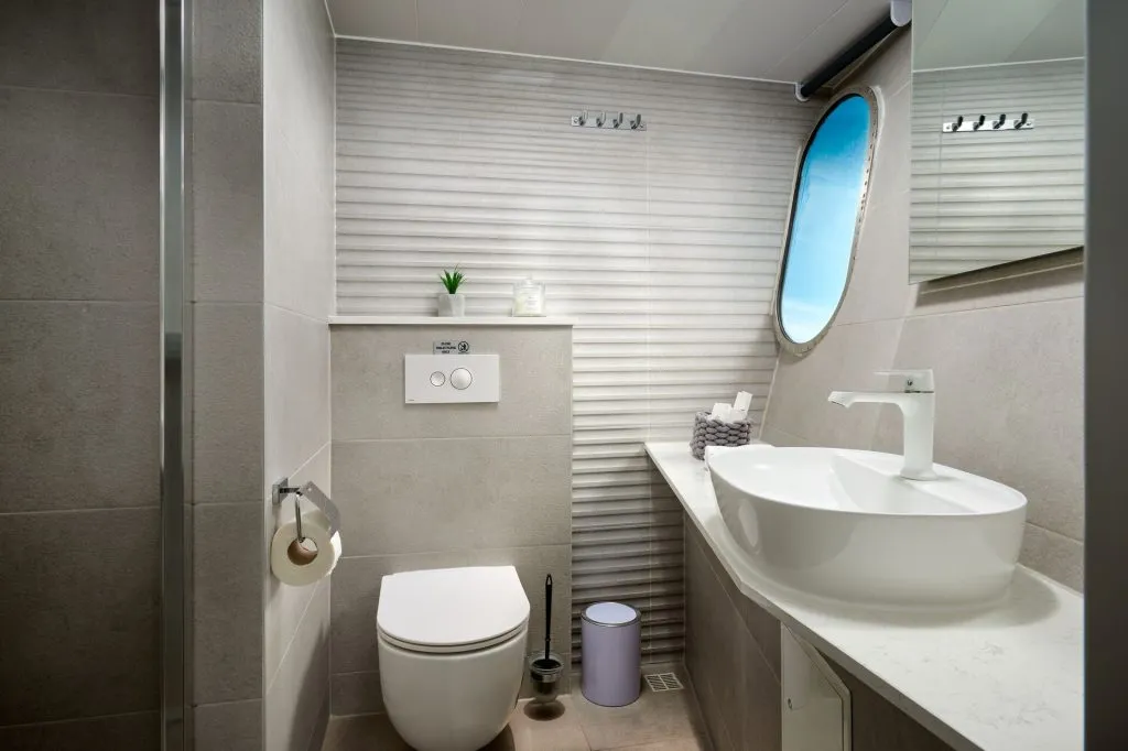 Modern bathroom with grey tiles, a wall-mounted toilet, white sink, round mirror, small plant, and a window with a blue sky view. Accessories include a toilet brush, waste bin, and toiletries on the counter.