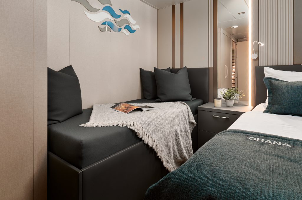 A cozy, modern bedroom with a small bed and a built-in sofa, both with dark cushions. A light throw, a magazine, and a plant are on the sofa. Abstract wave art decorates the wall above.