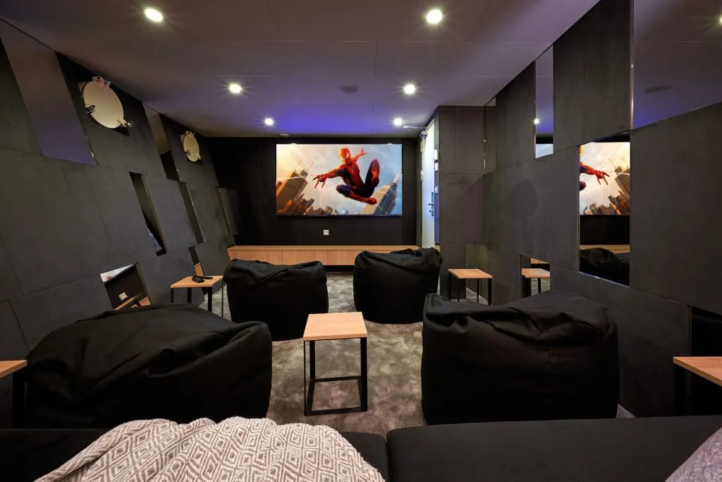 A modern home theater room with black bean bag chairs, small wooden side tables, and a large screen displaying Spider-Man. The walls are dark with reflective panels, and recessed ceiling lights illuminate the space.