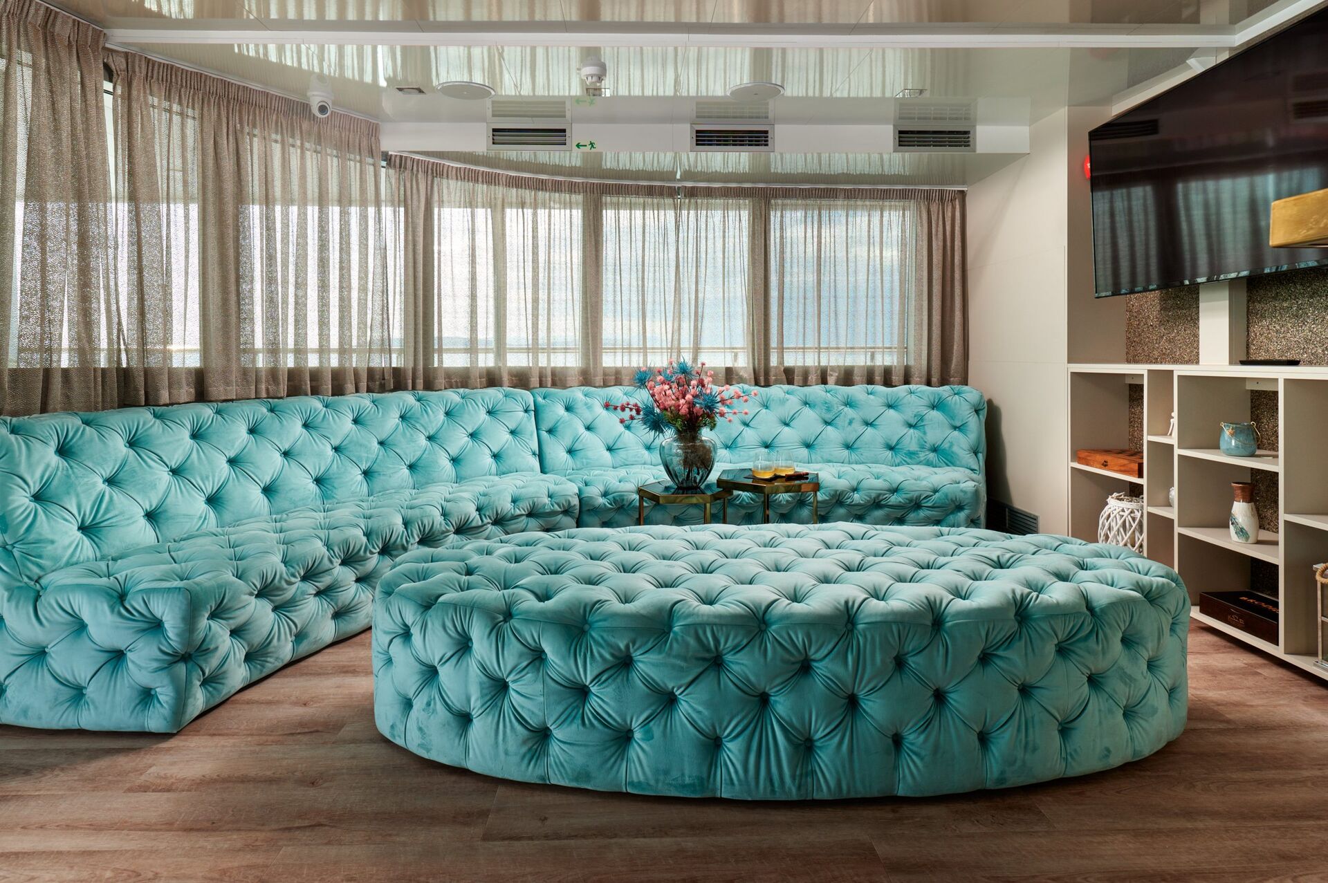 A spacious living room with a large, tufted turquoise sectional sofa and matching ottoman, sheer curtains on the windows, a small glass coffee table with a flower vase, and shelves with decorative items.