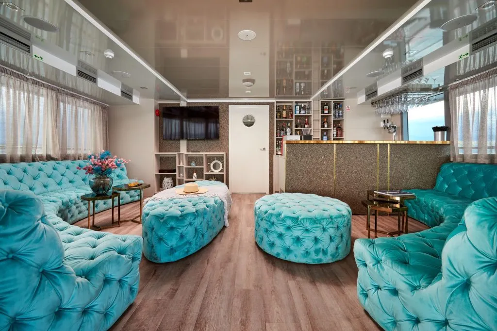 A stylish lounge with turquoise tufted sofas and ottomans, a gold-accented bar, wood flooring, and a wall-mounted TV. Large windows with sheer curtains provide natural light.