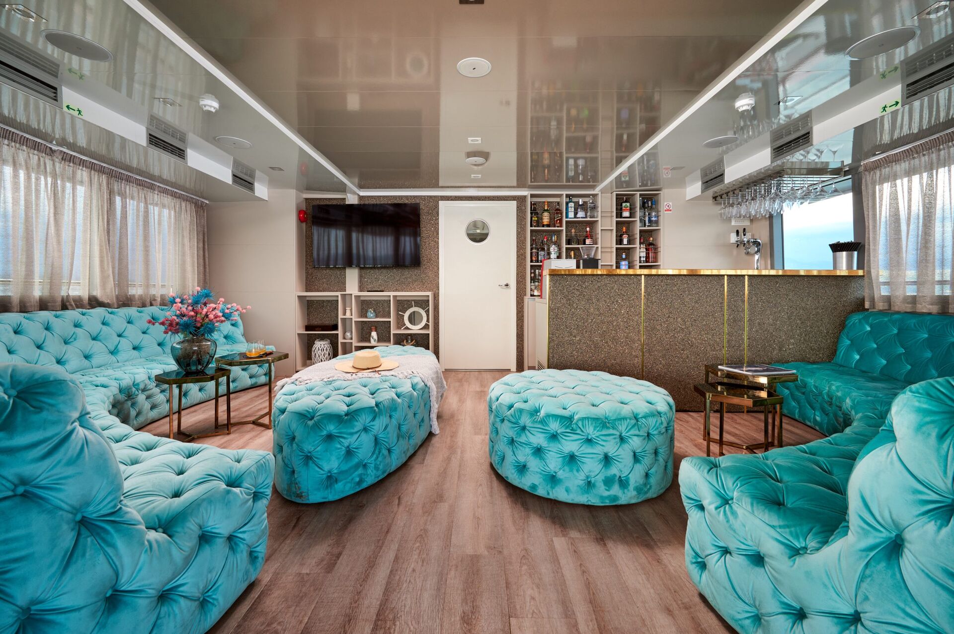 A stylish lounge with turquoise tufted sofas and ottomans, a gold-accented bar, wood flooring, and a wall-mounted TV. Large windows with sheer curtains provide natural light.