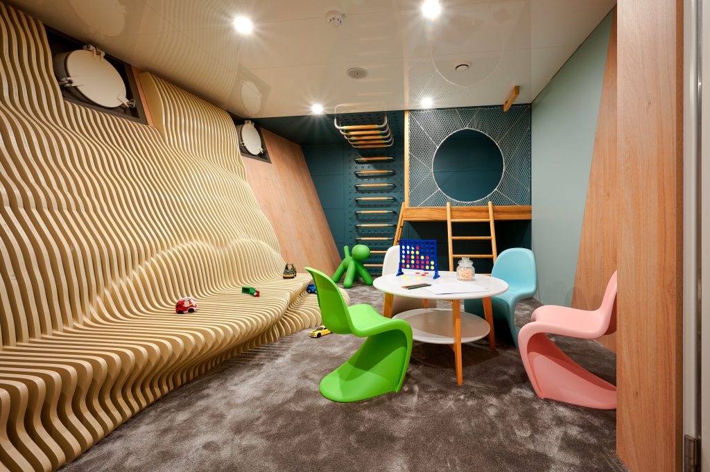 A modern playroom with a wavy wooden wall, climbing ladder, small round table with colorful chairs, toys on the floor, and soft gray carpet. Playful décor includes a green dog-shaped figure and a blue wall with a circular opening.