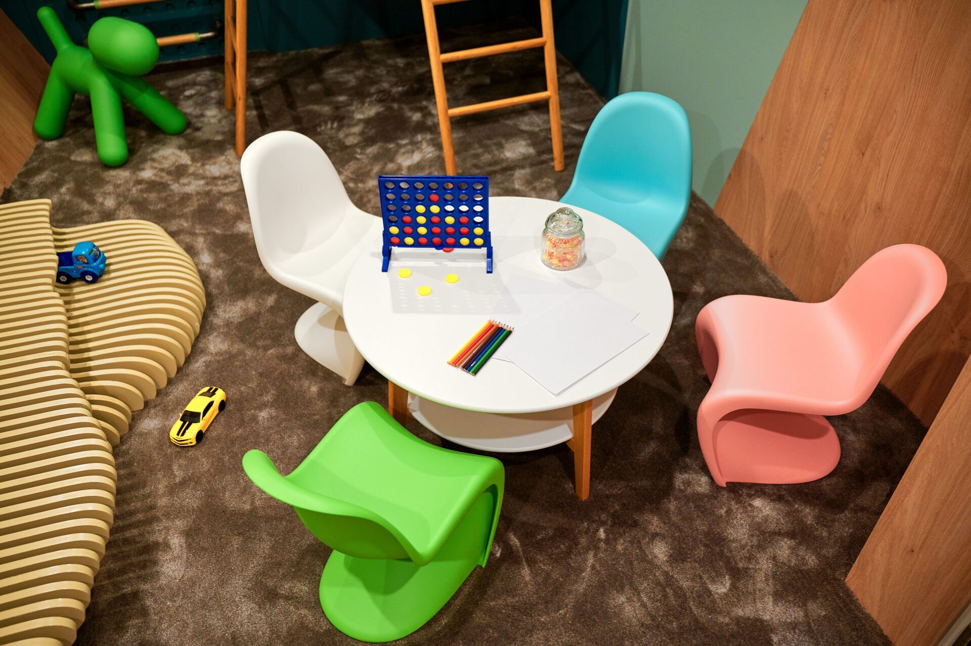 A round white table with colored chairs sits on a carpet, surrounded by toys, colored pencils, paper, a Connect Four game, and a jar of candy; a striped cushion and other play items are nearby in a cozy playroom.
