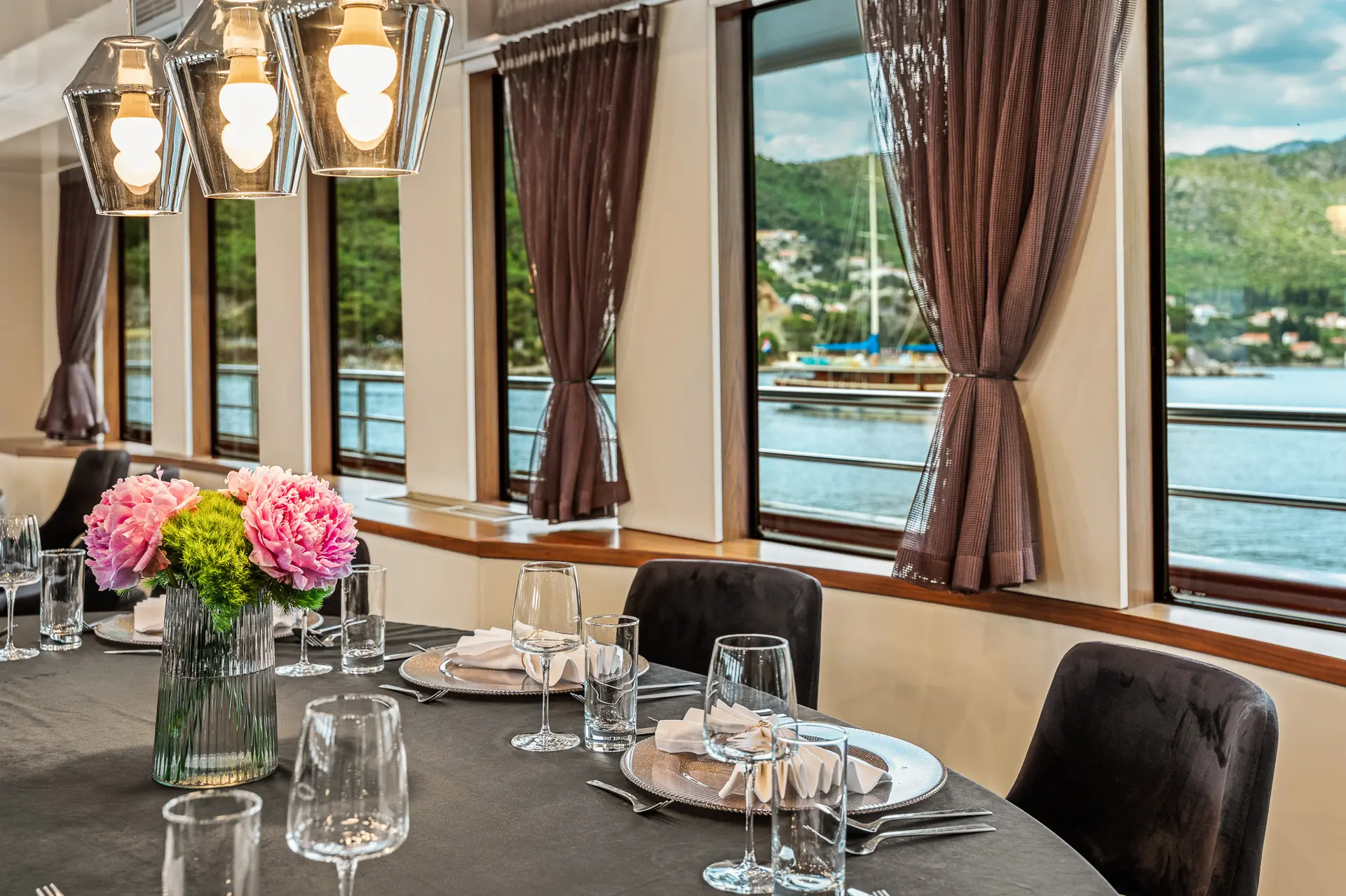 A round dining table set with glassware, plates, and a vase of pink flowers sits by large windows with brown curtains, offering a view of water, boats, and green hills outside. Modern pendant lights hang above the table.